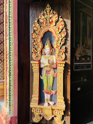 A decorative statue set within an ornate alcove, featuring a figure adorned with intricate details and vibrant colors. The alcove is richly carved with floral and ornamental patterns, highlighted by a bright yellow and red color scheme.