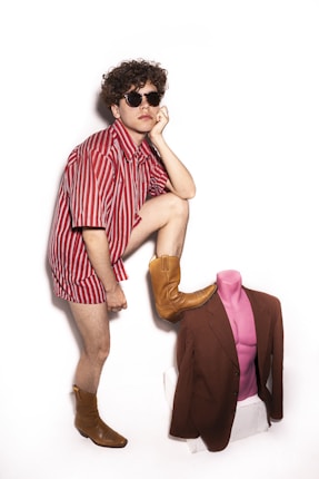 A person wearing an oversized red and white striped shirt and brown cowboy boots is standing with one leg resting on a pink mannequin torso dressed in a brown jacket. The individual is wearing dark sunglasses and has curly hair.