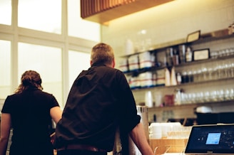 Two people standing at a counter in what appears to be a cafe or restaurant. Shelves lined with glassware and containers are visible in the background. A digital cash register or computer screen is on the counter.