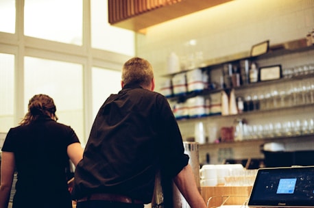 Two people standing at a counter in what appears to be a cafe or restaurant. Shelves lined with glassware and containers are visible in the background. A digital cash register or computer screen is on the counter.