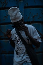 A person wearing dark sunglasses and a light-colored beanie stands against a weathered blue metal background. The T-shirt they are wearing features bold, graphic designs, including stylized faces and an aggressive animal. A dark bag is slung across their torso, and the overall lighting is dim, creating a mysterious and edgy atmosphere.