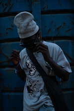 A person wearing dark sunglasses and a light-colored beanie stands against a weathered blue metal background. The T-shirt they are wearing features bold, graphic designs, including stylized faces and an aggressive animal. A dark bag is slung across their torso, and the overall lighting is dim, creating a mysterious and edgy atmosphere.