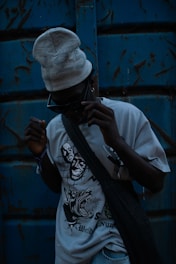 A person wearing dark sunglasses and a light-colored beanie stands against a weathered blue metal background. The T-shirt they are wearing features bold, graphic designs, including stylized faces and an aggressive animal. A dark bag is slung across their torso, and the overall lighting is dim, creating a mysterious and edgy atmosphere.