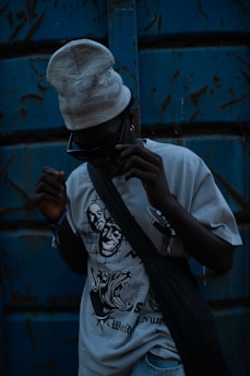 A person wearing dark sunglasses and a light-colored beanie stands against a weathered blue metal background. The T-shirt they are wearing features bold, graphic designs, including stylized faces and an aggressive animal. A dark bag is slung across their torso, and the overall lighting is dim, creating a mysterious and edgy atmosphere.