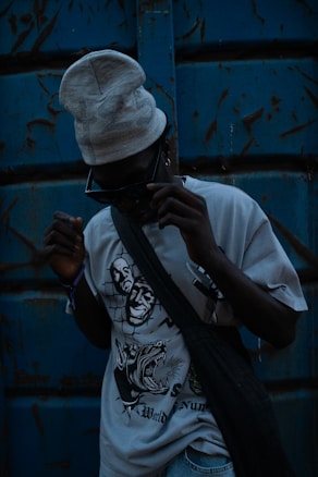 A person wearing dark sunglasses and a light-colored beanie stands against a weathered blue metal background. The T-shirt they are wearing features bold, graphic designs, including stylized faces and an aggressive animal. A dark bag is slung across their torso, and the overall lighting is dim, creating a mysterious and edgy atmosphere.