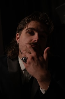A person is in semi-darkness, with their hand raised to their face, showcasing a large, ornate ring. The lighting casts dramatic shadows, creating a mysterious atmosphere. The individual has long hair and is dressed in a dark suit jacket, paired with a collared shirt.