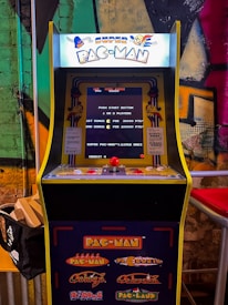 A vintage arcade game machine with the title 'Super Pac-Man' displayed prominently at the top. The machine features a screen with game instructions and scores, along with a joystick and buttons for gameplay. The background has a colorful graffiti-style wall with vibrant greens, oranges, and pinks.