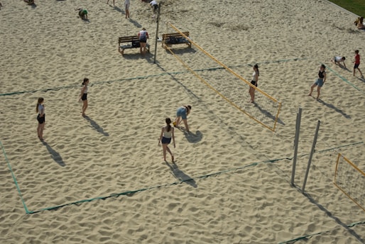 Several people are playing beach volleyball on sandy courts. The scene captures an active game with players positioned on either side of a volleyball net. The area is surrounded by sand, and some benches are visible in the background.