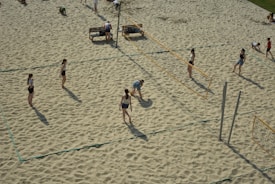 Several people are playing beach volleyball on sandy courts. The scene captures an active game with players positioned on either side of a volleyball net. The area is surrounded by sand, and some benches are visible in the background.
