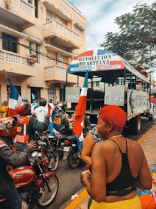 A street scene with a group of people on motorcycles, some holding flags, gathered near a truck with banners displaying 'Revolución Duartiana'. A woman with bright red hair stands in the foreground, surrounded by the vibrant colors of the event. The background features a multi-story building and trees.