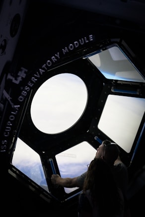 A person is looking out of a large circular window situated within a structure labeled 'ISS Cupola Observatory Module.' The view through the window appears to show space or a planetary surface below.