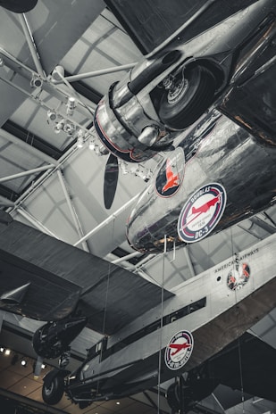 Several vintage airplanes are displayed in a museum setting with visible branding for American Airways and Douglas DC-3. The aircraft are suspended from the ceiling with visible structural elements, lighting fixtures, and metallic details.