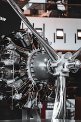 A close-up view of a radial aircraft engine displayed indoors. The complex machinery consists of numerous cylinders arranged in a circular pattern, with various metal rods and tubes connected to it. The background shows an industrial indoor space with some lighting fixtures and reflective surfaces.