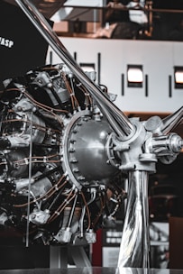 A close-up view of a radial aircraft engine displayed indoors. The complex machinery consists of numerous cylinders arranged in a circular pattern, with various metal rods and tubes connected to it. The background shows an industrial indoor space with some lighting fixtures and reflective surfaces.