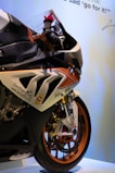A close-up view of a BMW sports motorcycle with detailed decals and labels on its body. The front wheel features metallic orange rims, and the motorcycle is positioned against a light background with writing and a signature visible at the top.