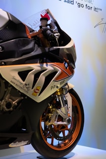 A close-up view of a BMW sports motorcycle with detailed decals and labels on its body. The front wheel features metallic orange rims, and the motorcycle is positioned against a light background with writing and a signature visible at the top.