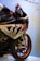 A close-up view of a BMW sports motorcycle with detailed decals and labels on its body. The front wheel features metallic orange rims, and the motorcycle is positioned against a light background with writing and a signature visible at the top.
