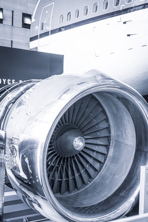 A close-up view of a large aircraft engine, prominently displaying the Rolls-Royce logo. The engine's fan blades and metallic exterior are visible, reflecting light. A portion of the aircraft's fuselage with windows is seen in the background.