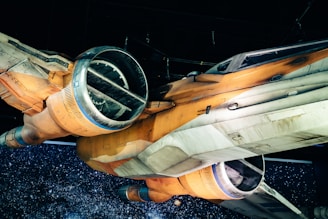 A detailed view of a sci-fi spacecraft with an orange and white body. The design features large circular thrusters and angular wings. The background is a starry space scene, enhancing the futuristic theme of the image.