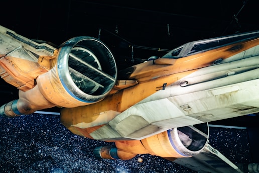 A detailed view of a sci-fi spacecraft with an orange and white body. The design features large circular thrusters and angular wings. The background is a starry space scene, enhancing the futuristic theme of the image.