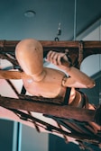 A wooden mannequin appears suspended between ropes attached to a rustic, wooden contraption. The mannequin is positioned horizontally, facing downward, showcasing a variety of joints and bends typical of an artist's model. The setup conveys complexity with ropes and mechanical elements interwoven around the mannequin.