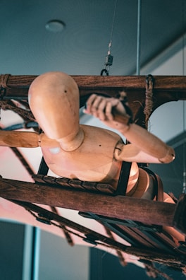 A wooden mannequin appears suspended between ropes attached to a rustic, wooden contraption. The mannequin is positioned horizontally, facing downward, showcasing a variety of joints and bends typical of an artist's model. The setup conveys complexity with ropes and mechanical elements interwoven around the mannequin.