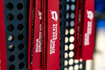 Red lanyards with the text 'National Air and Space Museum' hang against a perforated black metal backdrop. The text is printed in white and features a logo with a simple abstract design.