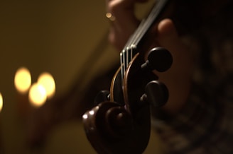 A close-up of a violin being played by a person, with soft, warm lighting creating a cozy and intimate atmosphere. The background is blurred, emphasizing the violin's pegs and fingerboard.