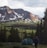 A scenic outdoor landscape with a lush green valley surrounded by tall pine trees. In the background, majestic mountains with patches of snow rise under a partially cloudy sky. In the foreground, a person wearing a red hat is walking near a green camping tent, creating a serene and peaceful camping scene.