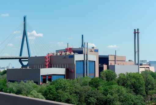 A large industrial building with a modern design and several tall smokestacks is surrounded by greenery. In the background, a cable-stayed bridge spans across the scene under a clear blue sky.