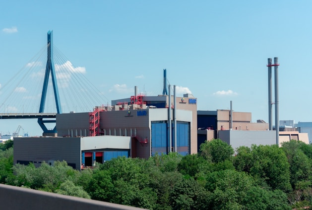 A large industrial building with a modern design and several tall smokestacks is surrounded by greenery. In the background, a cable-stayed bridge spans across the scene under a clear blue sky.