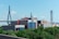 A large industrial building with a modern design and several tall smokestacks is surrounded by greenery. In the background, a cable-stayed bridge spans across the scene under a clear blue sky.