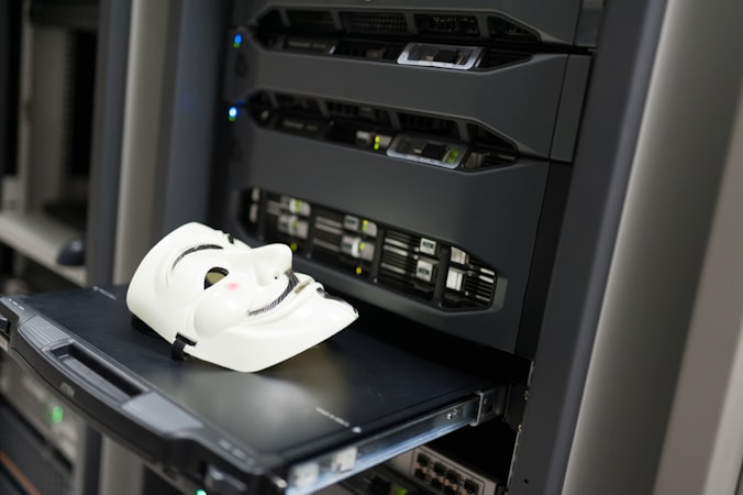 A white mask with distinct black features is placed on an open server tray in a data center or server room. The server is equipped with various LED indicators and network ports, suggesting an active setup.