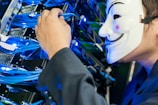 A person wearing a white mask is interacting with a network server, handling blue cables. The setup is illuminated with blue lights, suggesting a technological environment.