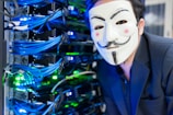 A person wearing a Guy Fawkes mask is positioned near a server rack filled with blue and green cables and illuminated lights. The individual is dressed in a dark-colored suit and is close to the camera.