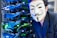 A person wearing a Guy Fawkes mask is positioned near a server rack filled with blue and green cables and illuminated lights. The individual is dressed in a dark-colored suit and is close to the camera.