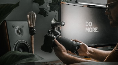A person is holding a camera with a large lens in a dimly lit room. A computer monitor displaying the message 'DO MORE.' is positioned in the background. Surrounding elements include a speaker and some plants, enhancing the ambiance.