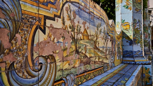 A series of intricately painted ceramic tiles displaying a landscape scene with faded artistic depictions. The tiles are adorned with patterns and floral motifs, showcasing a combination of blues, yellows, greens, and browns. Some portions of the artwork appear aged and worn, revealing a sense of history.