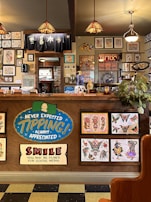 The image depicts the interior of a tattoo parlor. Artwork of various tattoo designs is displayed on the walls and a wooden counter. The counter features colorful signs related to tipping and social media. There are pendant lights with stained glass shades hanging from the ceiling, and a variety of framed pictures and designs provide a vibrant backdrop. A water cooler is present on the counter, and a collection of plants adds a touch of greenery.