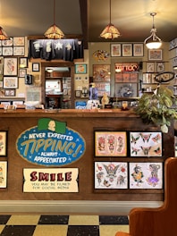 The image depicts the interior of a tattoo parlor. Artwork of various tattoo designs is displayed on the walls and a wooden counter. The counter features colorful signs related to tipping and social media. There are pendant lights with stained glass shades hanging from the ceiling, and a variety of framed pictures and designs provide a vibrant backdrop. A water cooler is present on the counter, and a collection of plants adds a touch of greenery.