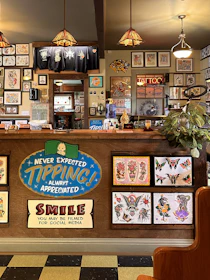 The image depicts the interior of a tattoo parlor. Artwork of various tattoo designs is displayed on the walls and a wooden counter. The counter features colorful signs related to tipping and social media. There are pendant lights with stained glass shades hanging from the ceiling, and a variety of framed pictures and designs provide a vibrant backdrop. A water cooler is present on the counter, and a collection of plants adds a touch of greenery.