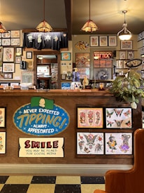 The image depicts the interior of a tattoo parlor. Artwork of various tattoo designs is displayed on the walls and a wooden counter. The counter features colorful signs related to tipping and social media. There are pendant lights with stained glass shades hanging from the ceiling, and a variety of framed pictures and designs provide a vibrant backdrop. A water cooler is present on the counter, and a collection of plants adds a touch of greenery.