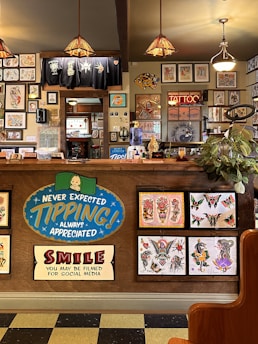 The image depicts the interior of a tattoo parlor. Artwork of various tattoo designs is displayed on the walls and a wooden counter. The counter features colorful signs related to tipping and social media. There are pendant lights with stained glass shades hanging from the ceiling, and a variety of framed pictures and designs provide a vibrant backdrop. A water cooler is present on the counter, and a collection of plants adds a touch of greenery.