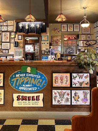 The image depicts the interior of a tattoo parlor. Artwork of various tattoo designs is displayed on the walls and a wooden counter. The counter features colorful signs related to tipping and social media. There are pendant lights with stained glass shades hanging from the ceiling, and a variety of framed pictures and designs provide a vibrant backdrop. A water cooler is present on the counter, and a collection of plants adds a touch of greenery.