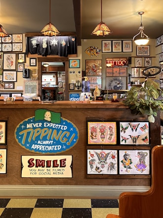 The image depicts the interior of a tattoo parlor. Artwork of various tattoo designs is displayed on the walls and a wooden counter. The counter features colorful signs related to tipping and social media. There are pendant lights with stained glass shades hanging from the ceiling, and a variety of framed pictures and designs provide a vibrant backdrop. A water cooler is present on the counter, and a collection of plants adds a touch of greenery.