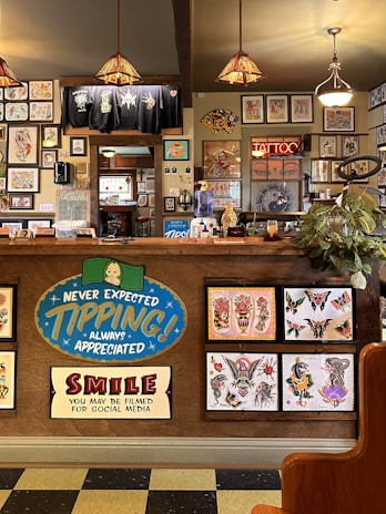 The image depicts the interior of a tattoo parlor. Artwork of various tattoo designs is displayed on the walls and a wooden counter. The counter features colorful signs related to tipping and social media. There are pendant lights with stained glass shades hanging from the ceiling, and a variety of framed pictures and designs provide a vibrant backdrop. A water cooler is present on the counter, and a collection of plants adds a touch of greenery.