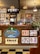 The image depicts the interior of a tattoo parlor. Artwork of various tattoo designs is displayed on the walls and a wooden counter. The counter features colorful signs related to tipping and social media. There are pendant lights with stained glass shades hanging from the ceiling, and a variety of framed pictures and designs provide a vibrant backdrop. A water cooler is present on the counter, and a collection of plants adds a touch of greenery.