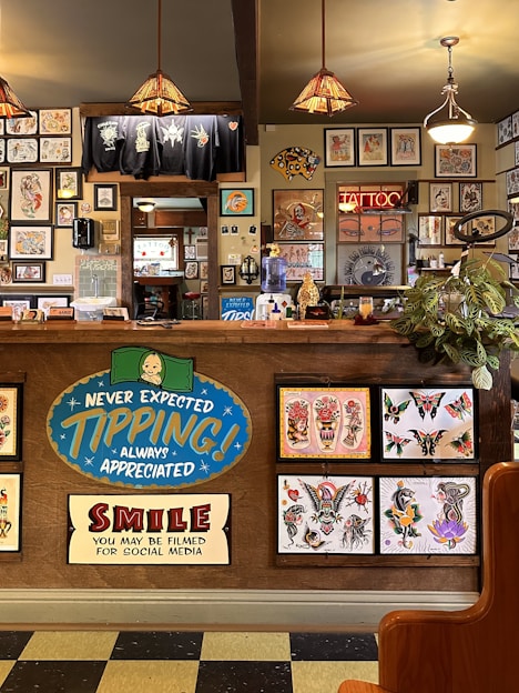 The image depicts the interior of a tattoo parlor. Artwork of various tattoo designs is displayed on the walls and a wooden counter. The counter features colorful signs related to tipping and social media. There are pendant lights with stained glass shades hanging from the ceiling, and a variety of framed pictures and designs provide a vibrant backdrop. A water cooler is present on the counter, and a collection of plants adds a touch of greenery.