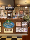 The image depicts the interior of a tattoo parlor. Artwork of various tattoo designs is displayed on the walls and a wooden counter. The counter features colorful signs related to tipping and social media. There are pendant lights with stained glass shades hanging from the ceiling, and a variety of framed pictures and designs provide a vibrant backdrop. A water cooler is present on the counter, and a collection of plants adds a touch of greenery.