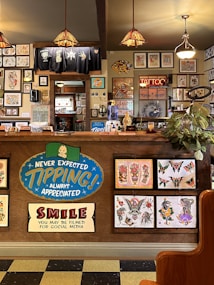 The image depicts the interior of a tattoo parlor. Artwork of various tattoo designs is displayed on the walls and a wooden counter. The counter features colorful signs related to tipping and social media. There are pendant lights with stained glass shades hanging from the ceiling, and a variety of framed pictures and designs provide a vibrant backdrop. A water cooler is present on the counter, and a collection of plants adds a touch of greenery.